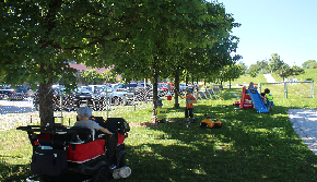 Das Bild zeigt einen eingezäunten Garten mit grüner Wiese und Bäumen. Im Vordergrund steht ein Krippenbus mit 4 Plätzen. Im Hintergrund ist eine blau/rote Rutsche zu sehen. Im Garten spielen mehrere Kinder.