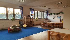 Das Bild zeigt den Kindergarten, mit einem blauem Teppich auf dem ein Globus leuchtet und eine Zugstation aus Holz zu sehen ist. Im Hintergrund befindet sich ein großer Kindergartentisch mit 10 Sitzplätzen.