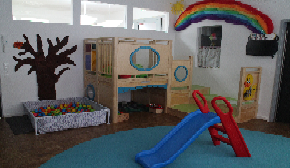 Das Bild zeigt einen Gruppenraum, mit einem blauem Teppich auf dem eine Rutsche steht und im Hintergrund befindet sich eine Hochebene über dem ein Regenbogen hängt.