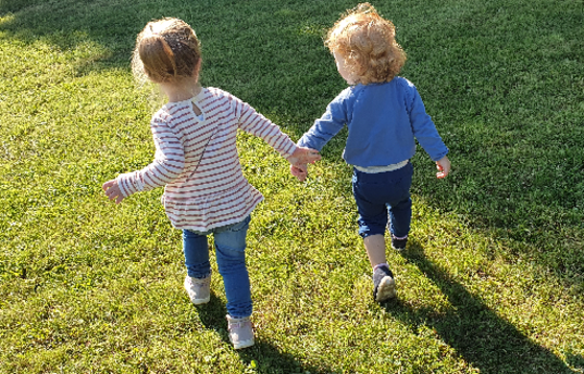 Auf dem Bild ist zu sehen, dass zwei Krippenkinder, welche sich an der Hand fassen, über eine grüne Wiese laufen.